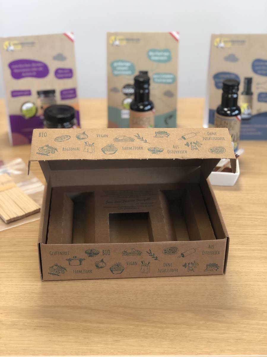 Leere Geschenkbox des Stillen Verkostungs-Sets auf dem Tisch, dahinter stehen die Produkte samt Aufsteller und Verkostungspipetten. 