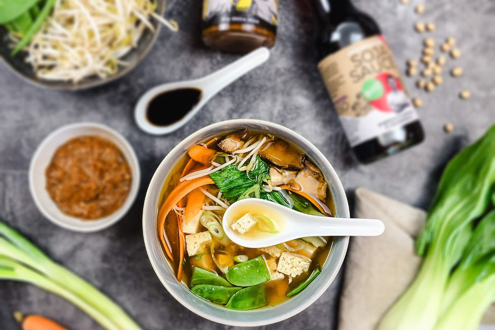 Flatlay-Aufnahme: Schale heißer Miso-Suppe mit Tofu, Frühlingszwiebeln & Wakame, daneben Löffel, Misopaste und saisonales Gemüse. 